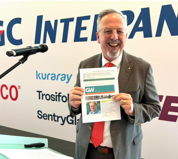 Smiling man in suit holds a printed document while standing at a podium with a microphone. - © Matthias Rehbgerger / GW