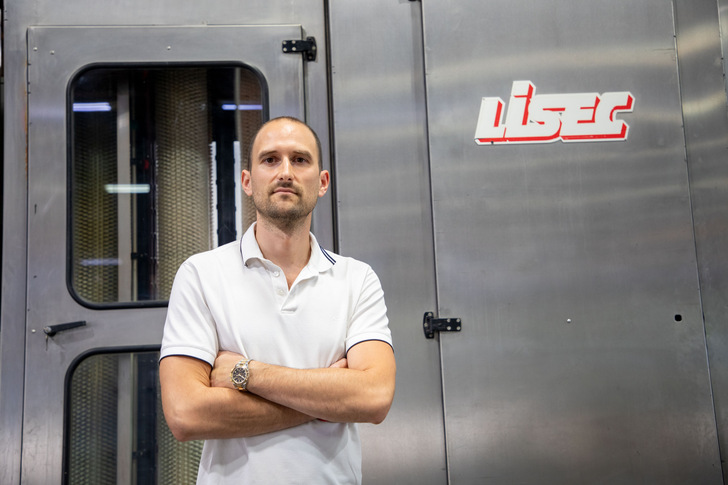 Man in white polo shirt stands with arms crossed in front of industrial machine with "laser" sign. - © Lisec