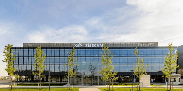 Modern glass building with the "Şişecam" logo, trees in front, and a clear blue sky above. - © Şişecam