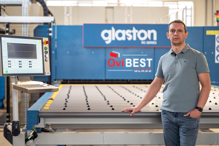 Ein Mann steht an einer Glasbearbeitungsmaschine in einer Fabrik, hinter ihm die Schilder "glaston" und "OviBEST". - © OviBest