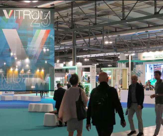 Man geht durch eine moderne überdachte Messehalle mit einem großen "VITRUM 25"-Schild und Messeständen. - © Vitrum