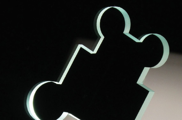 A silhouette of a puzzle piece with two round ends, illuminated from behind, against a dark background. - © Lisec