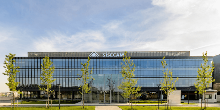 Modern glass building with "SISecam" logo, reflecting the sky, fronted by young trees and a clear, blue sky. - © Şişecam