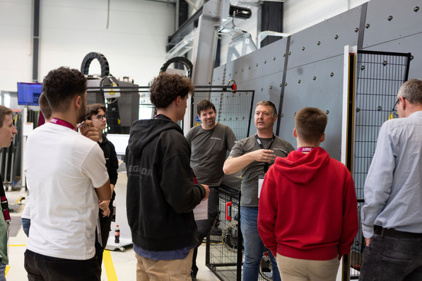 A group of people listens to a man explaining machinery in an industrial workshop setting. - © Lisec